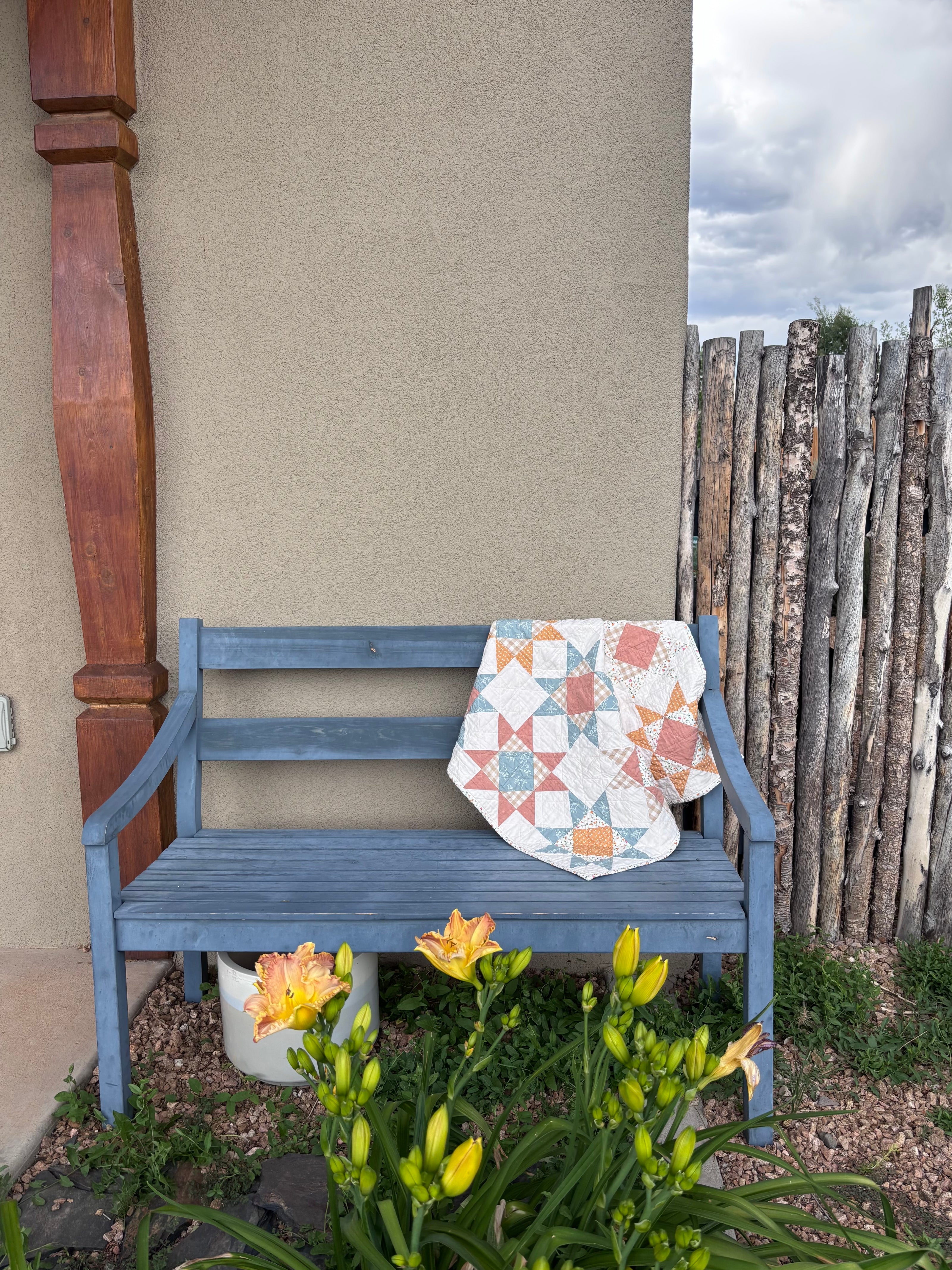 Blue bench with a pink and yellow baby quilt draped over it in a garden setting.