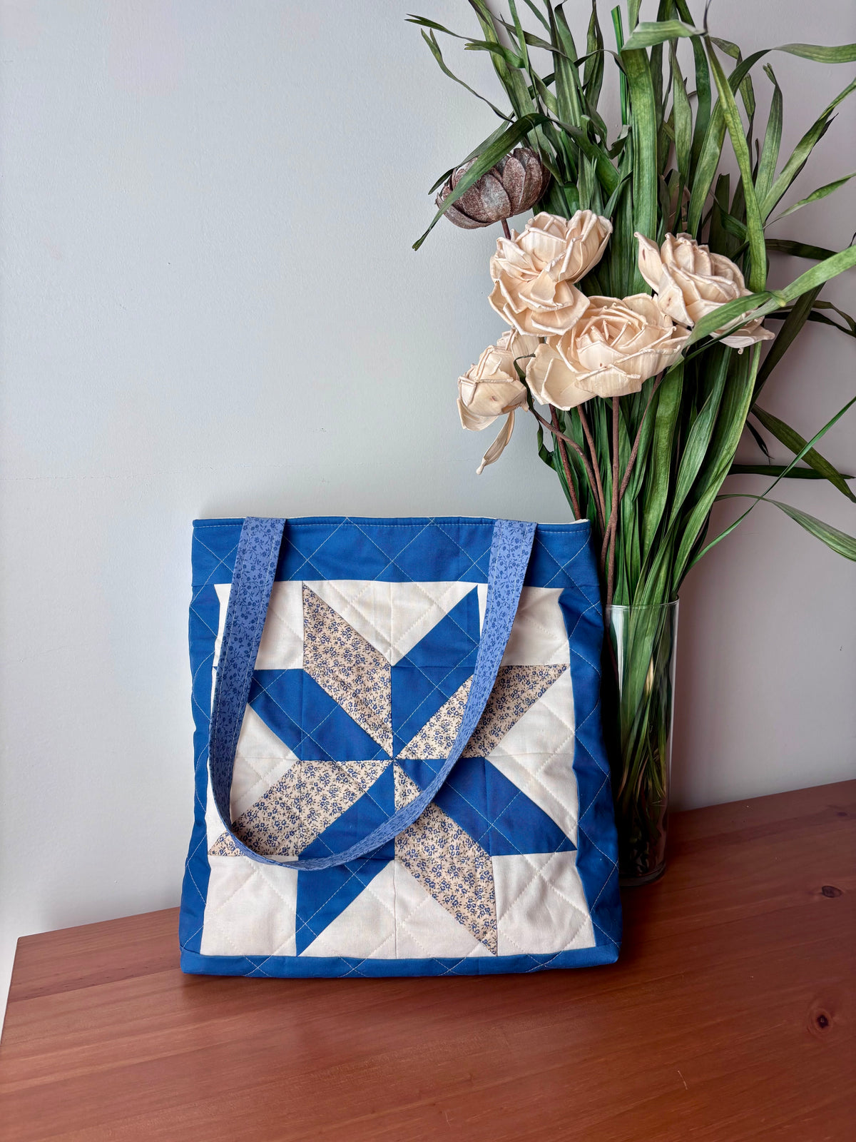 A quilted fabric tote bag with a blue and white starburst pattern sits on a wooden table next to a glass vase filled with dried flowers and green stems.