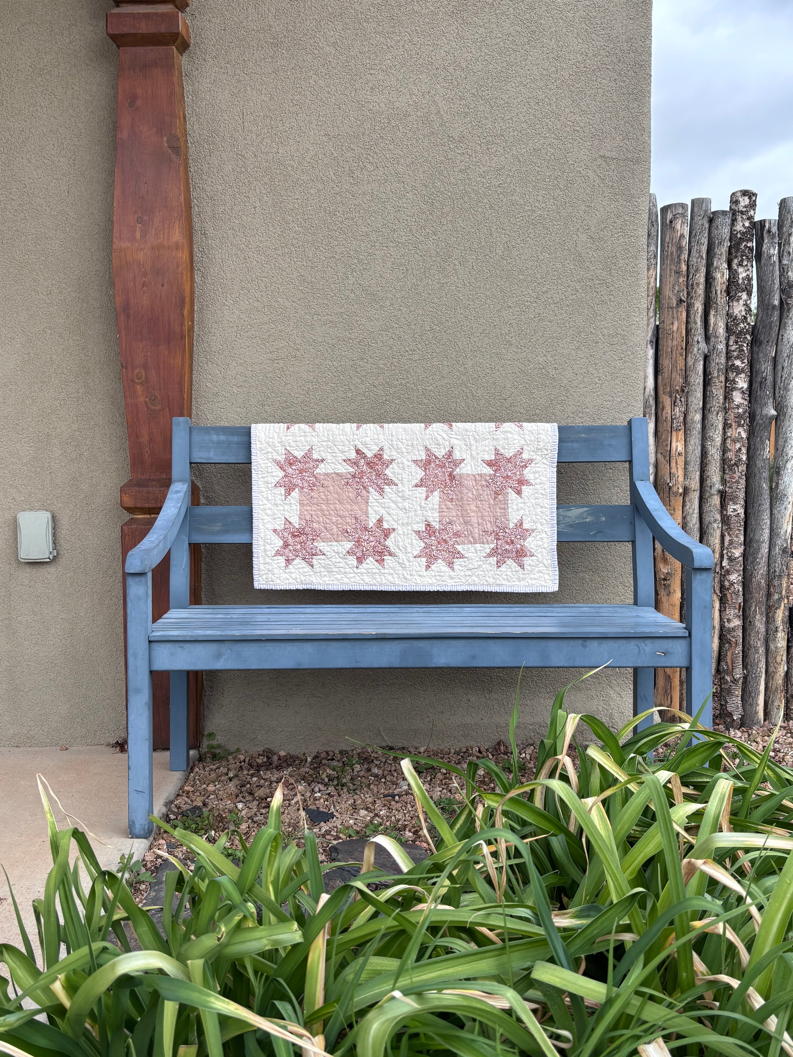 Blue bench with a quilt draped over it against a beige wall, surrounded by greenery.