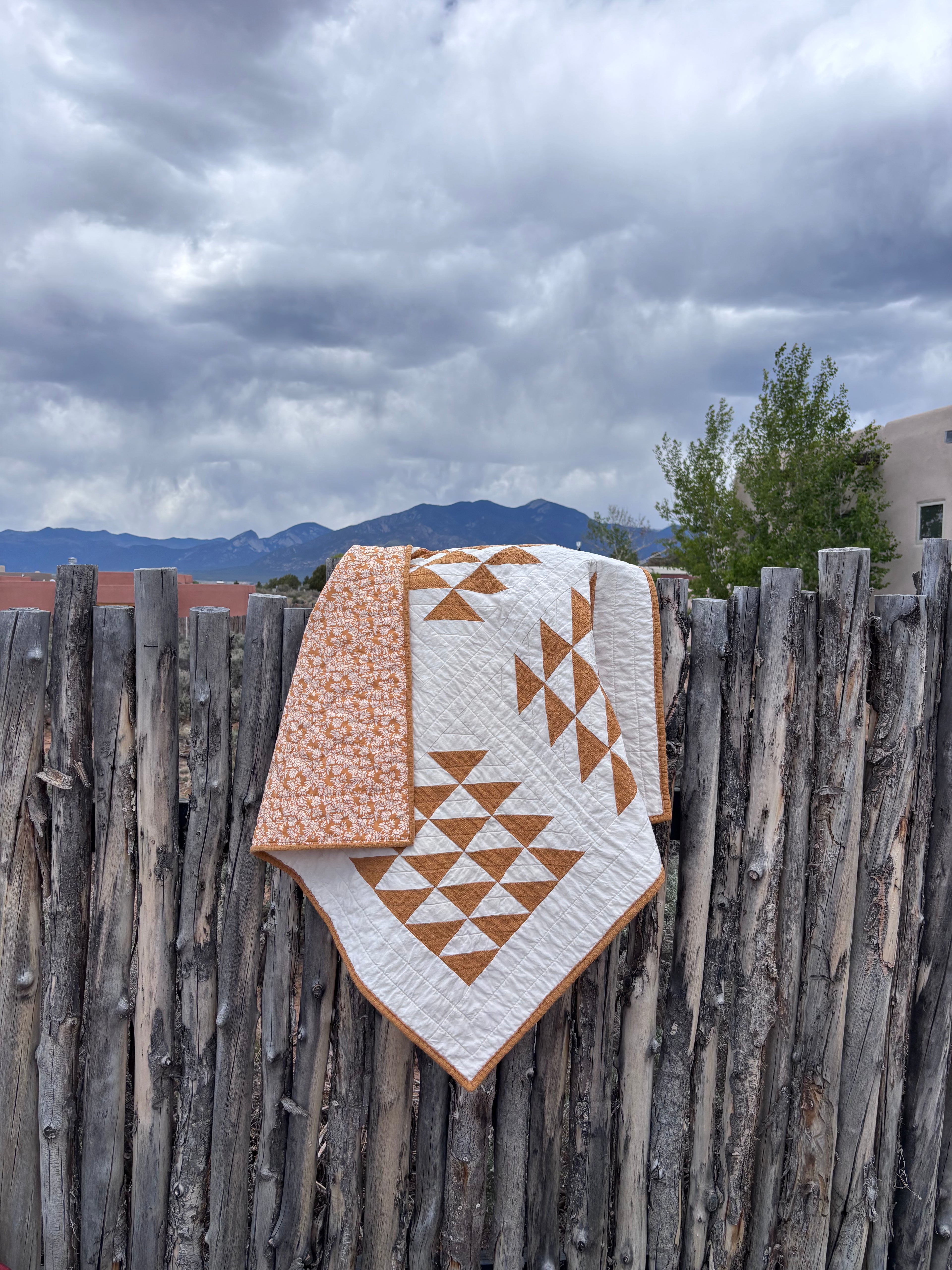 Quilt with geometric pattern draped over a wooden fence against a cloudy sky.